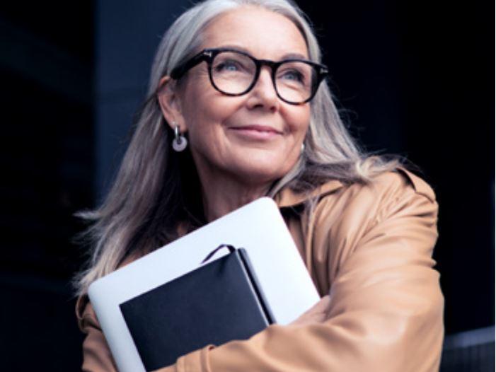 Uma imagem de uma mulher mais velha de óculos sorrindo e carregando um notebook.