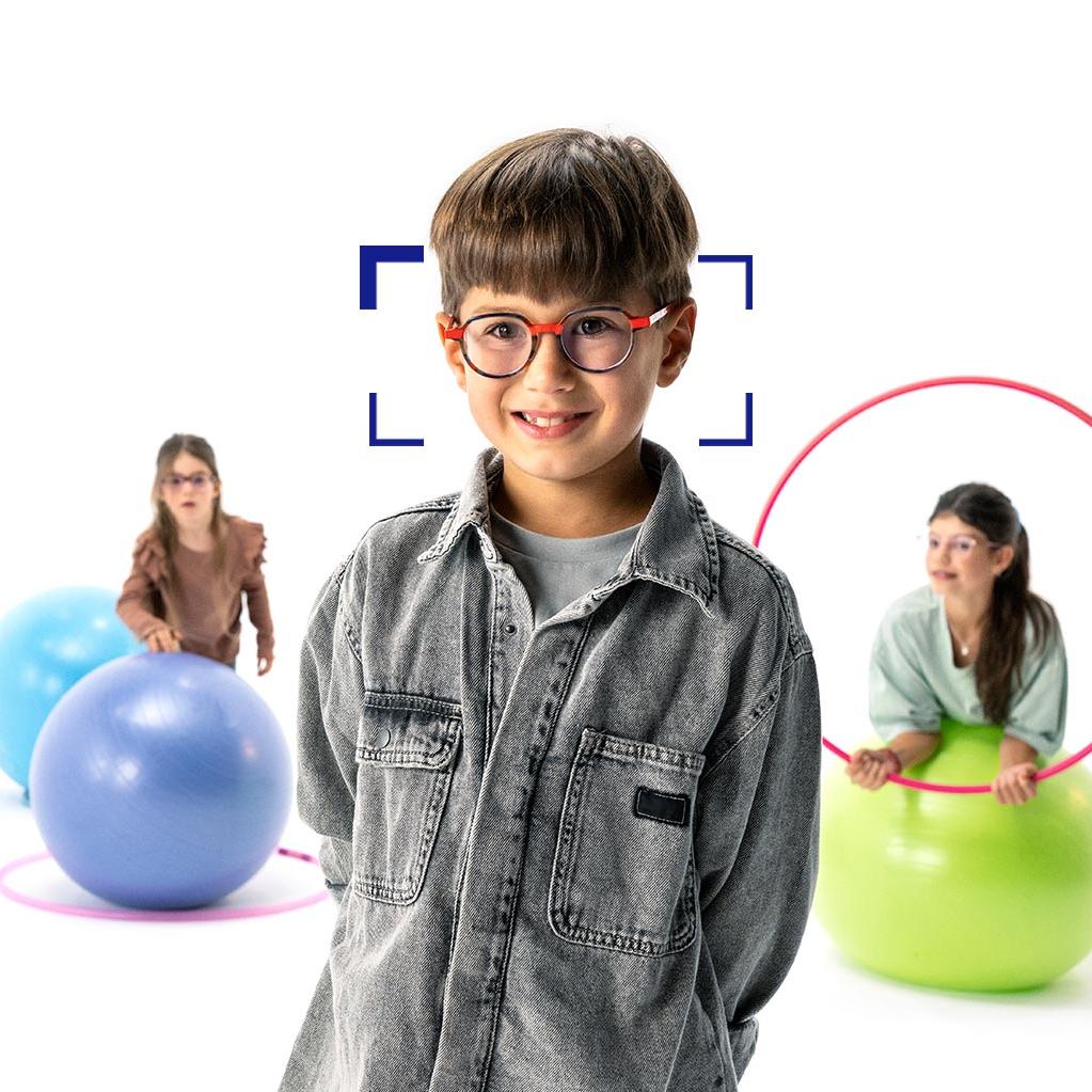 Menino de cabelos castanhos usando óculos redondos com lentes ZEISS MyoCare, posicionado em primeiro plano, sorrindo para a câmera. Ao fundo, duas meninas usando lentes ZEISS MyoCare brincam com aros e bolas de ginástica.