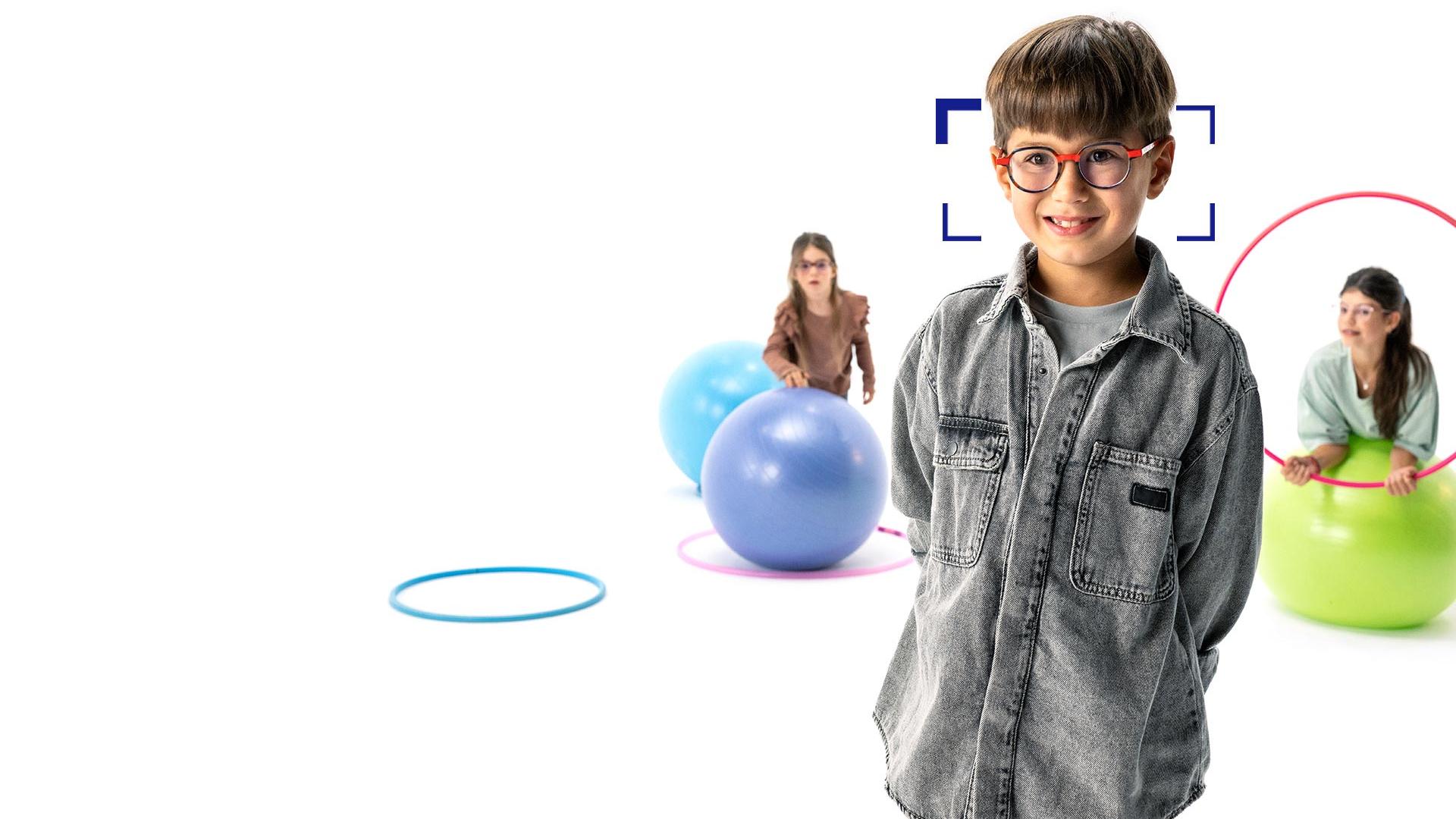 Menino de cabelos castanhos usando óculos redondos com lentes ZEISS MyoCare, posicionado em primeiro plano, sorrindo para a câmera. Ao fundo, duas meninas usando lentes ZEISS MyoCare brincam com aros e bolas de ginástica.
