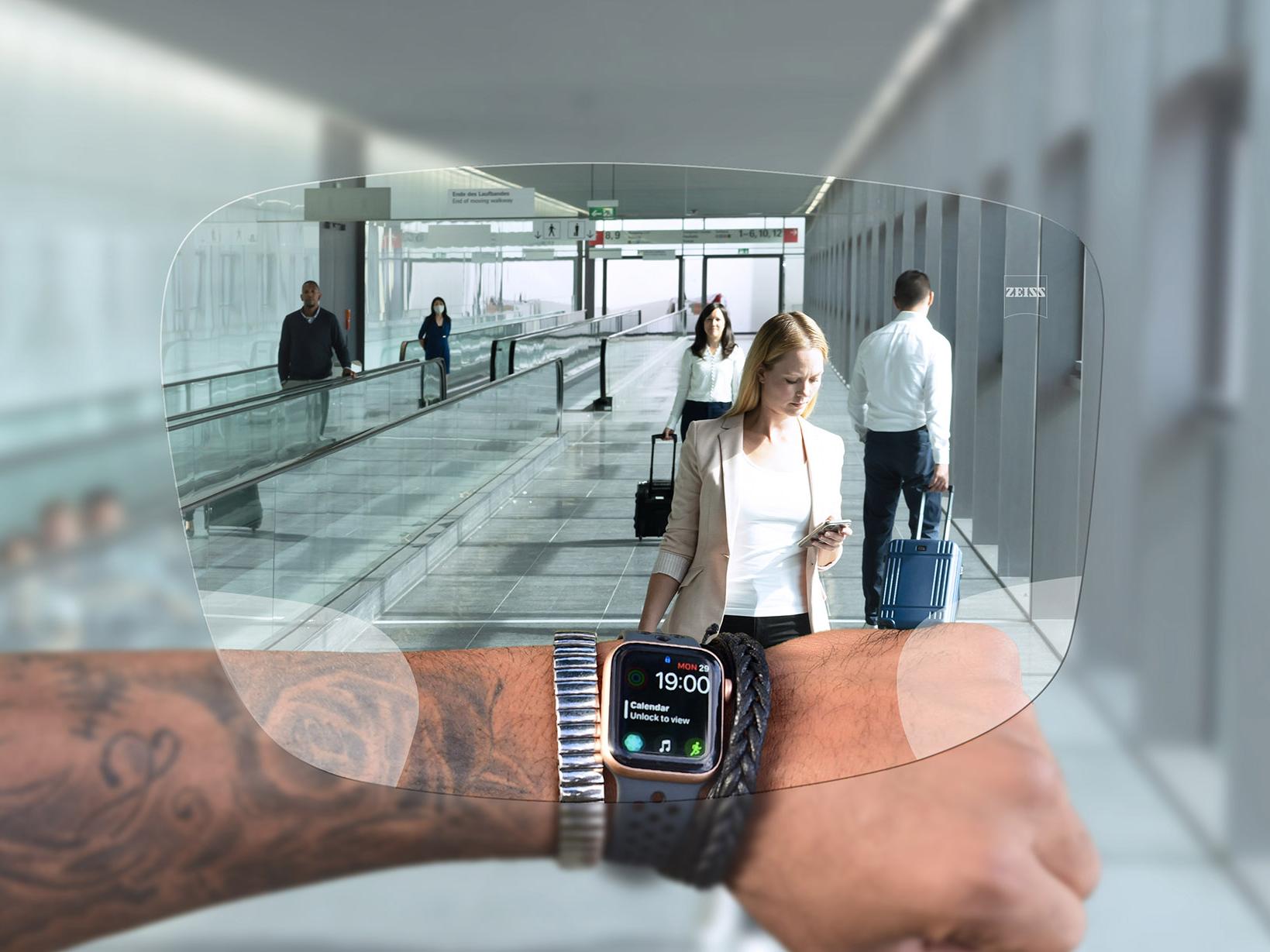 Pessoa olhando para o relógio através das lentes ZEISS SmartLife Digital. Ao fundo, vê-se uma situação típica de aeroporto, com pessoas carregando malas e andando nas escadas rolantes. 