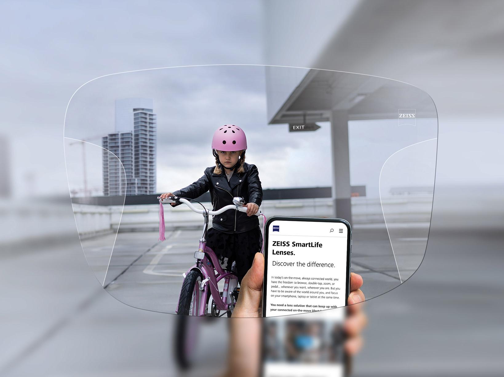 Uma pessoa olhando para o celular através das lentes ZEISS SmartLife Progressivas Ao fundo, vê-se uma menina de capacete cor-de-rosa andando de bicicleta. 