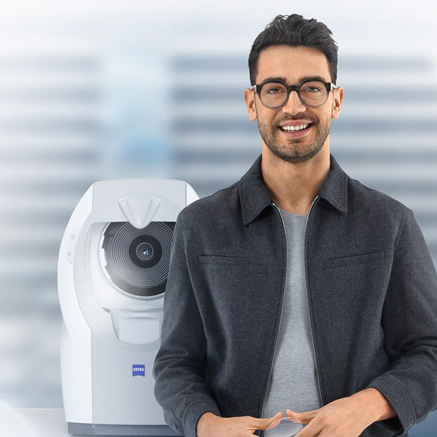 Um jovem de cabelos castanhos curtos, com lentes ZEISS, sorrindo para a câmera. Ao fundo, vê-se um ZEISS i.Profiler plus.