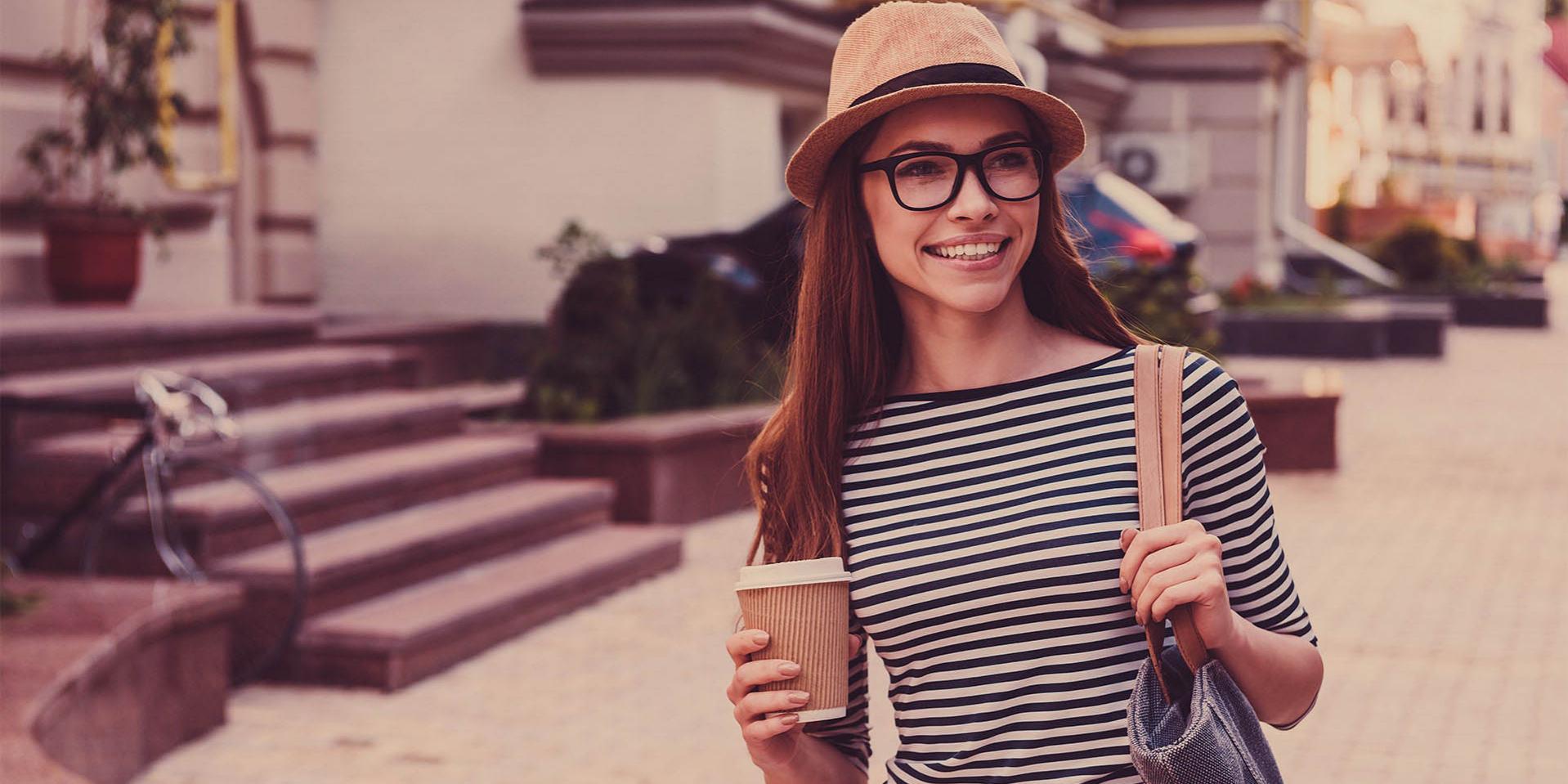 Uma jovem de boné e óculos com uma caneca de café na mão (à direita) e uma bolsa no ombro esquerdo