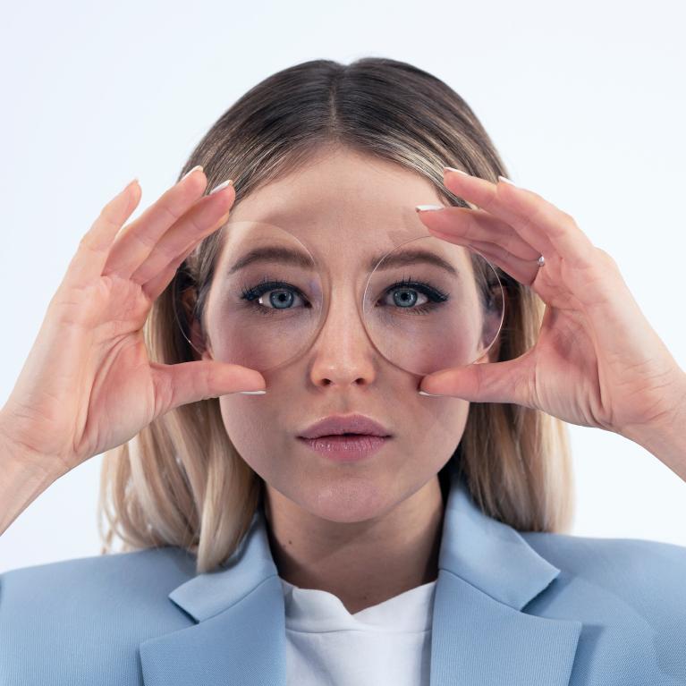 Mulher jovem e loira segurando lentes em frente aos olhos para mostrar a boa aparência, sem efeitos que distorcem os olhos, promovida pelas lentes de visão simples ZEISS ClearView.