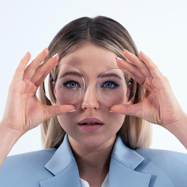 Mulher jovem e loira segurando lentes em frente aos olhos para mostrar o efeito de olhos minúsculos provocado por óculos grossos negativos.