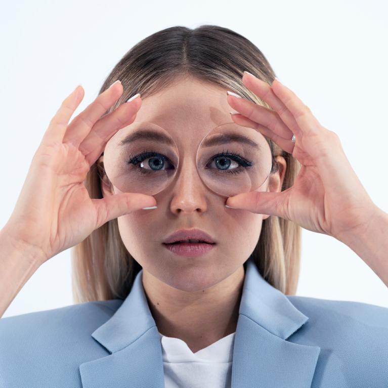 Mulher jovem e loira segurando lentes em frente aos olhos para mostrar o efeito de olho de peixe provocado por óculos grossos.