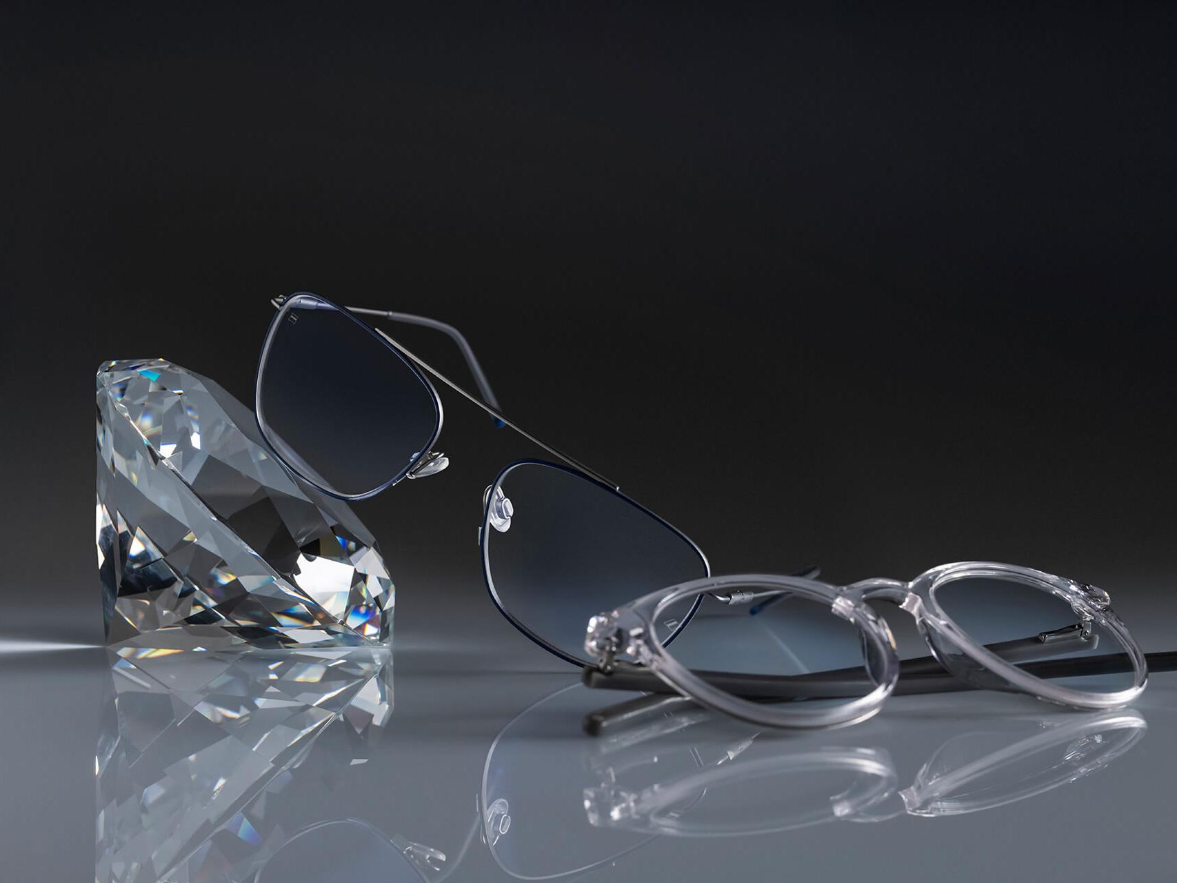 Dois pares de óculos com lentes ZEISS que têm o revestimento de lente mais duro possível – DuraVision® Platinum. Um par de óculos encostado em um cristal, o outro está no chão. Ambos têm lentes lentes incolores sem reflexo azulado.