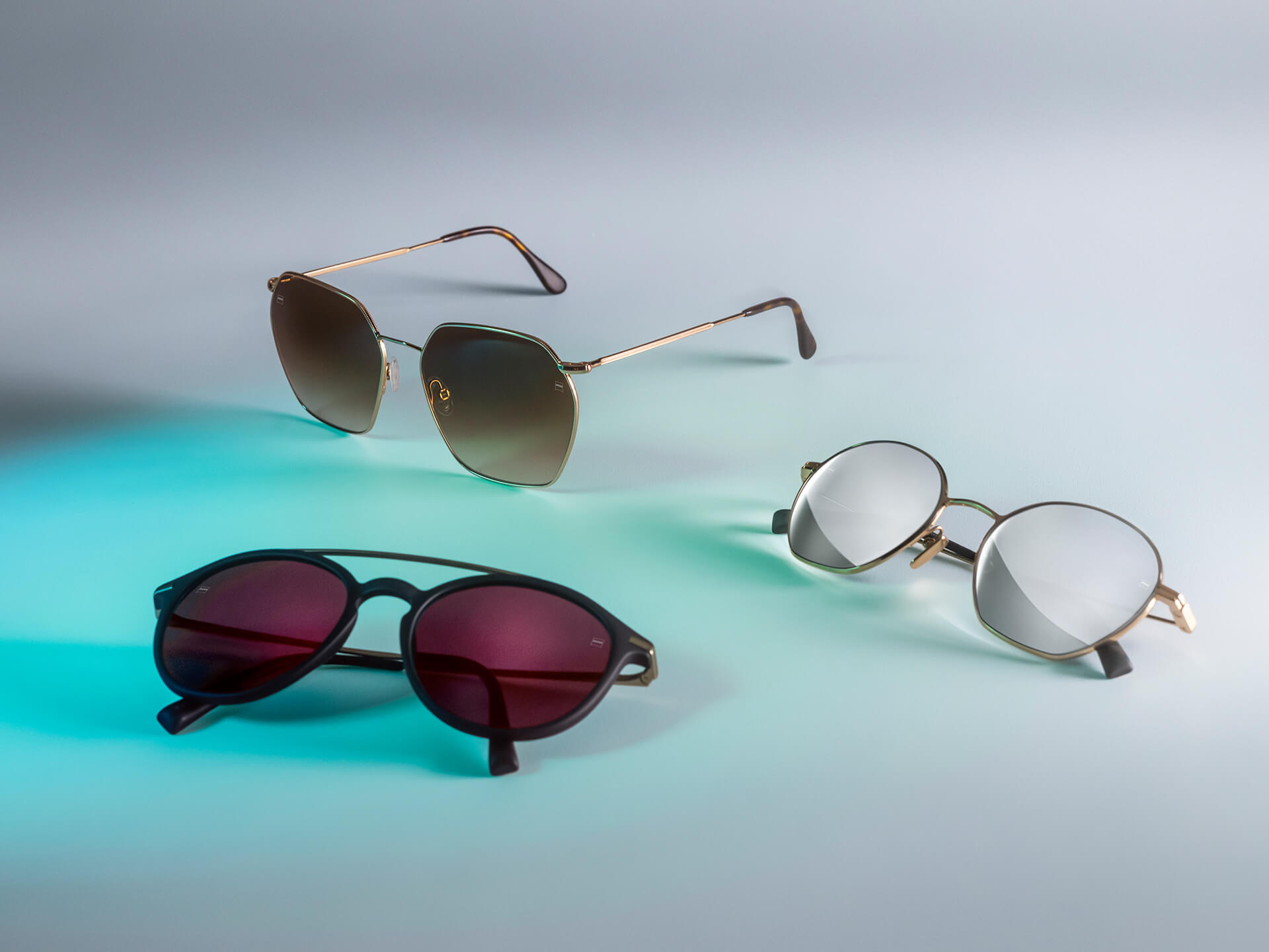 Três pares de óculos de sol com diferentes lentes coloridas para óculos de sol da ZEISS, contendo tratamentos DuraVision Sun, DuraVision Mirror e Flash Mirror, visíveis sobre um fundo branco com reflexo de luz azul.