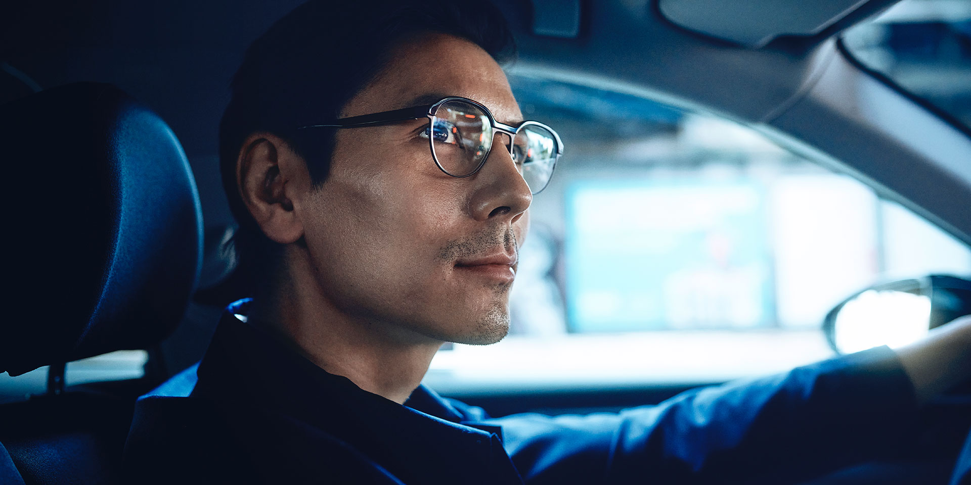 Homem dirigindo um carro, com um olhar confiante e um leve sorriso. Ele usa lentes de visão simples ZEISS DriveSafe.