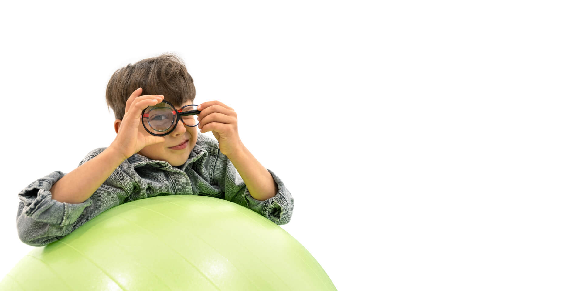 Menino usando lentes para gerenciamento da miopia, debruçado sobre uma bola de ginástica e segurando uma lupa na frente de um dos olhos.