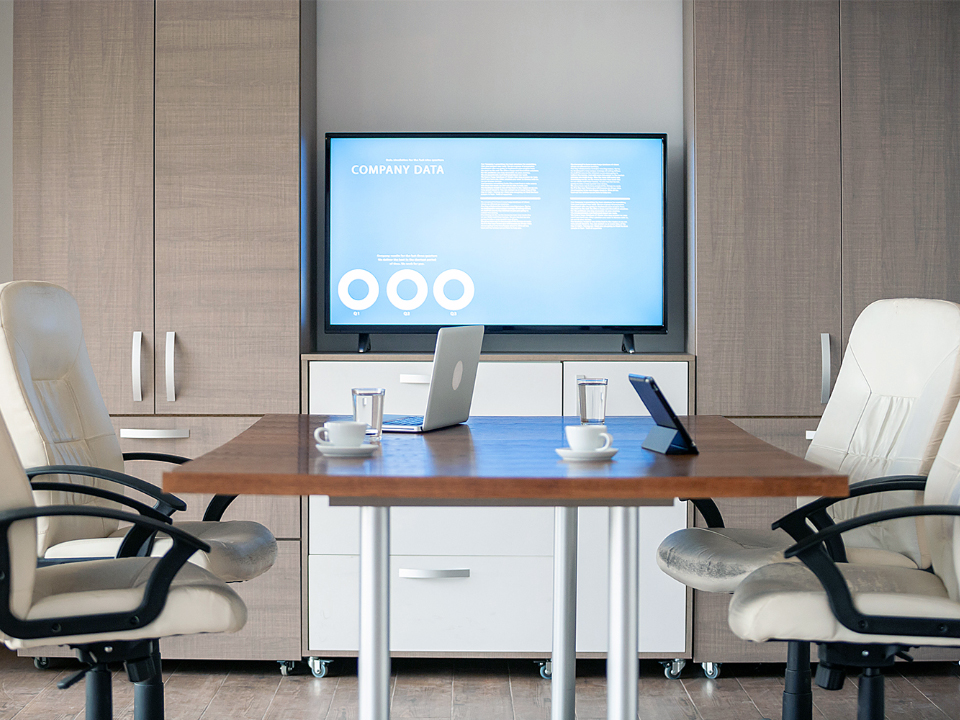 Visão da mesa de um escritório executivo. Um monitor está fixado à parede a alguns metros de distância. Há uma pequena mesa de reunião com cadeiras e laptops.