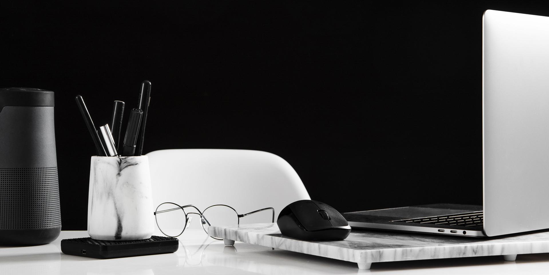 Local de trabalho organizado mostrando um laptop aberto e um mouse à dirieita. Na frente do laptop, há um par de óculos. À esquerda da imagem, há uma caixa com canetas.