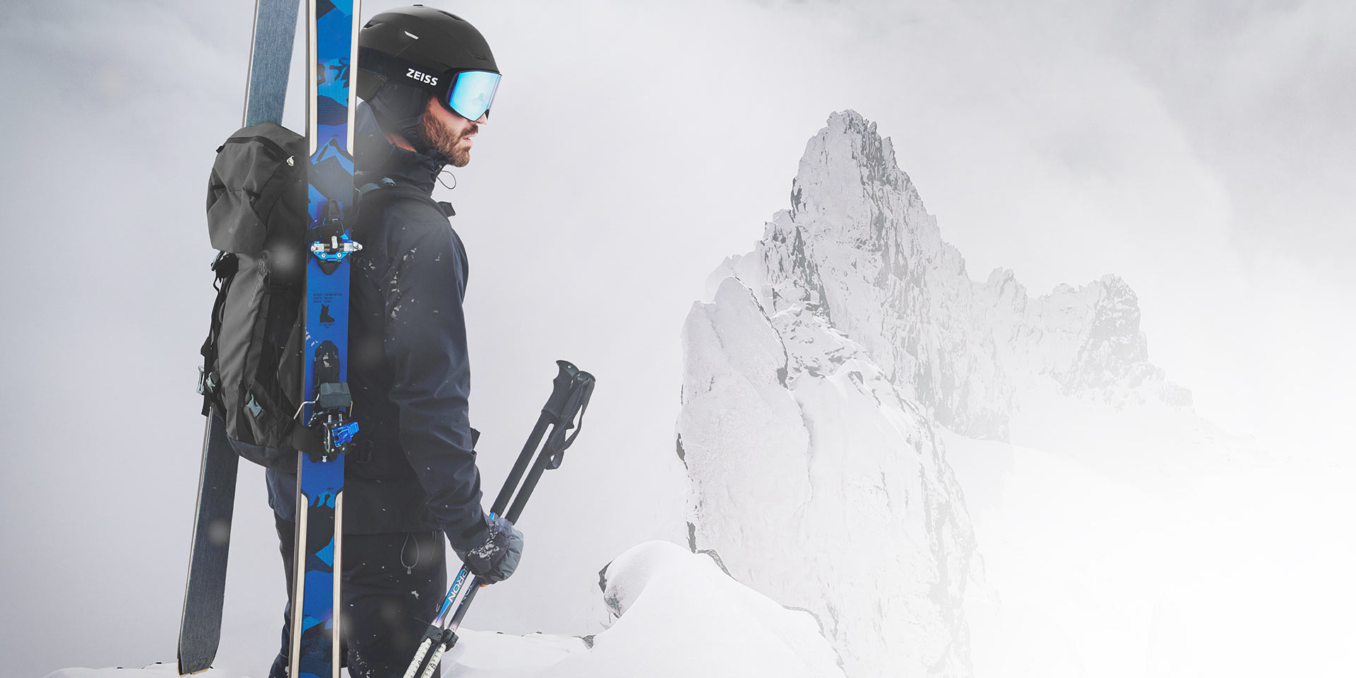 A man wears a black helmet with blue mirrored ZEISS ski goggles, has skis and a backpack on his back and holds the ski poles in his right hand. He is standing on a mountain top.