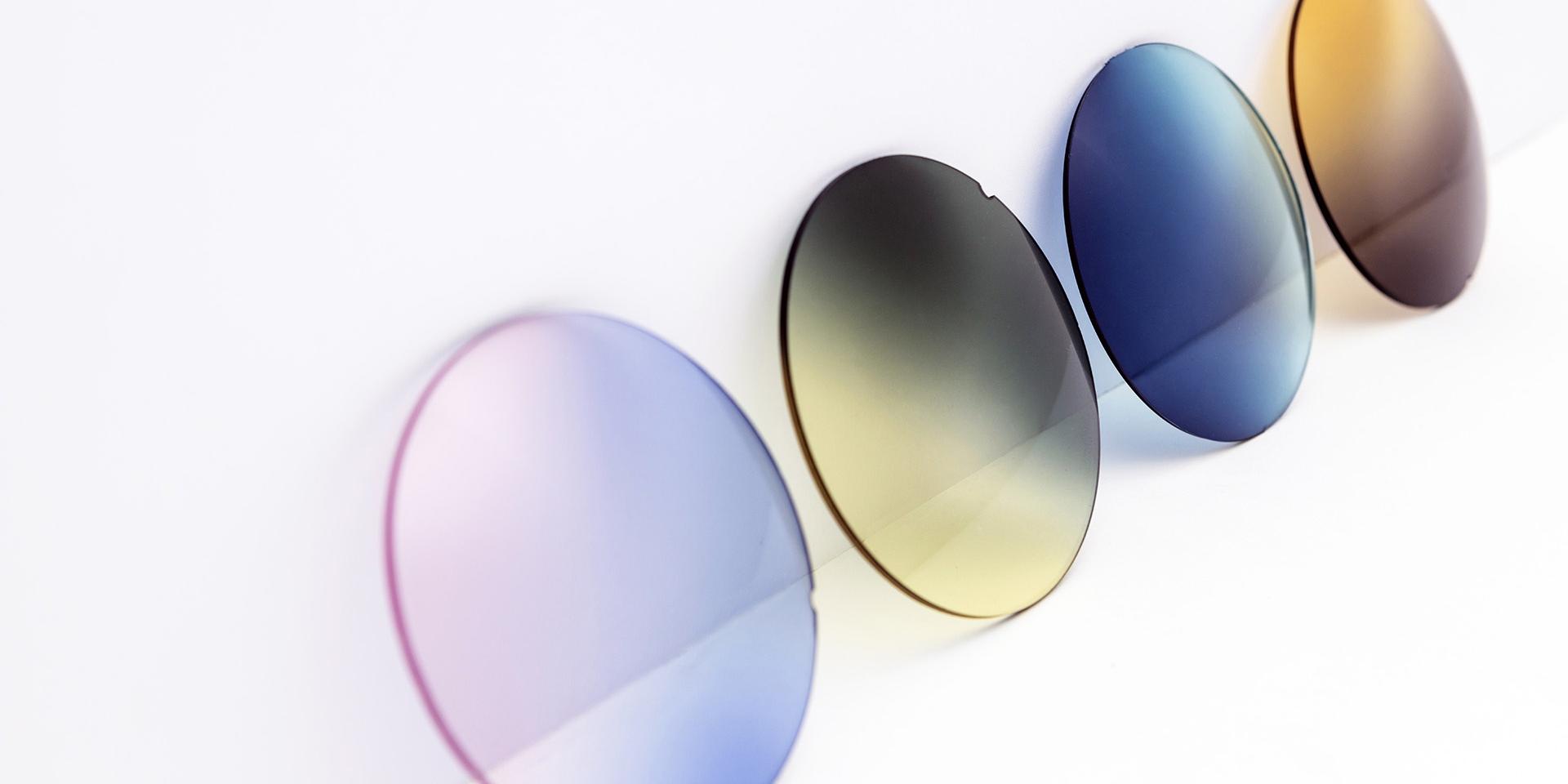 Diferentes lentes coloridas para óculos de sol sobre uma superfície branca: rosa arroxeado, cinza amarelado, azul e marrom.